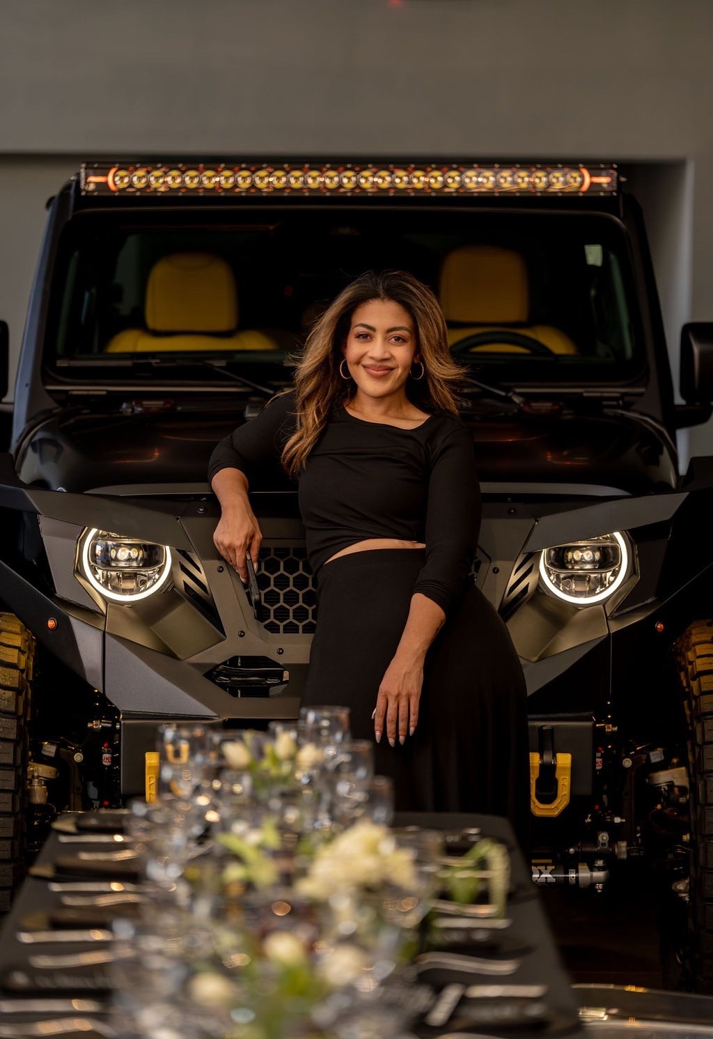 Alexandria Reed in black, leaning on a custom black Jeep with a set dinner table in the foreground